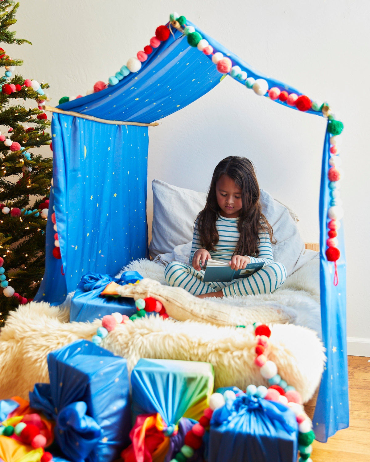 A child sitting inside a colorful Christmas canopy with Christmas Tree and gifts seen outside sarasilk
